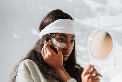 Young African American female in headband applying eye patch on face while looking in mirror on white background