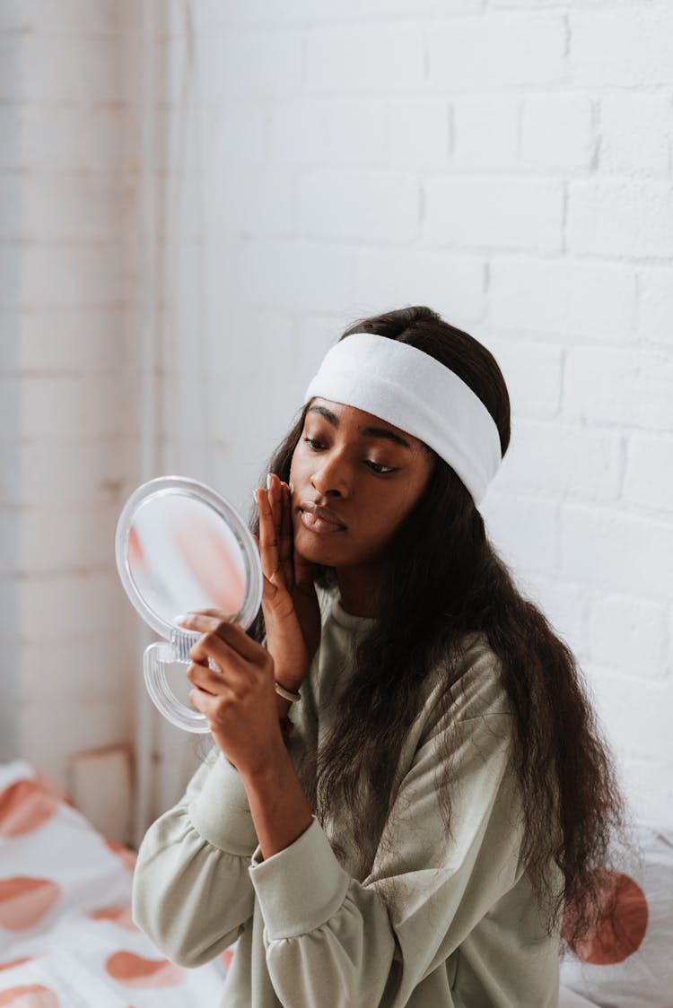 Black Woman Applying Hydrating Cream On Face Against Mirror Indoors