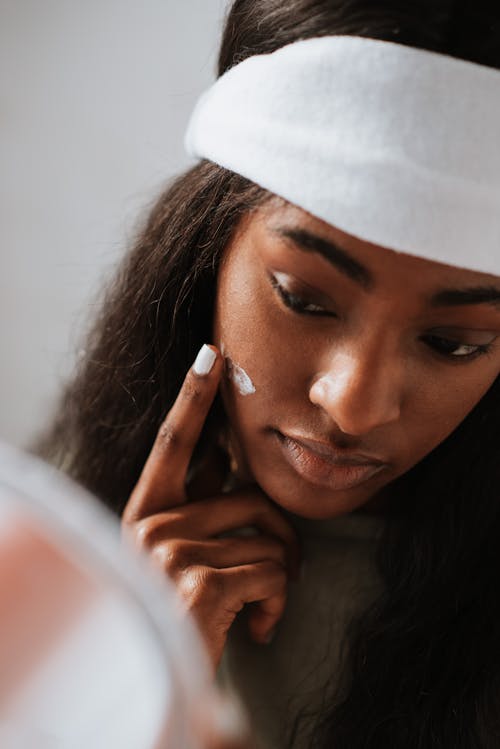 Crop young black female in headband applying hydrating cream on face against mirror on light background