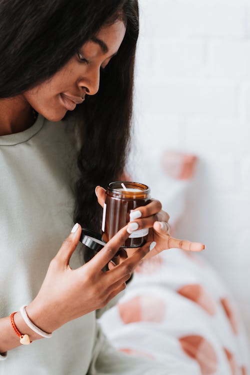 Crop young African American female with long hair holding jar with candle on bed at home