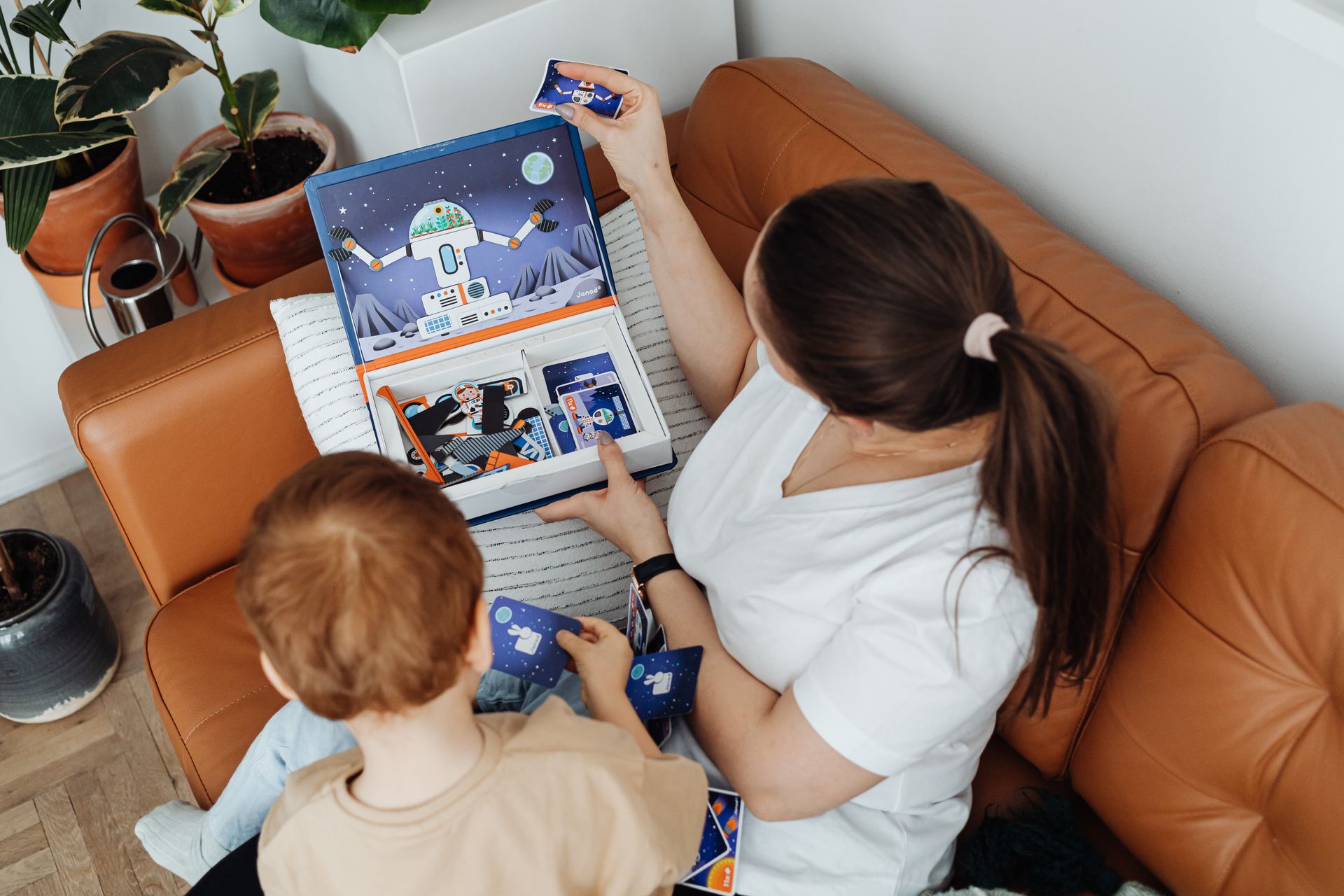 A mother and child playing an educational card game on a sofa, surrounded by houseplants.