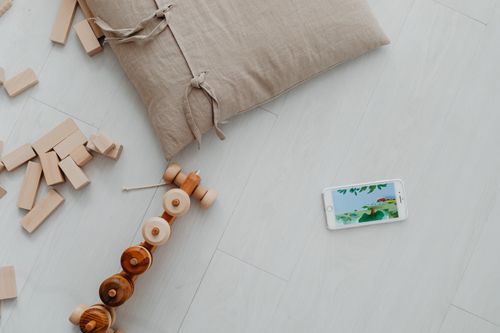 Free A flat lay of wooden toys, a smartphone, and a cushion on a light floor, offering a minimalist aesthetic. Stock Photo