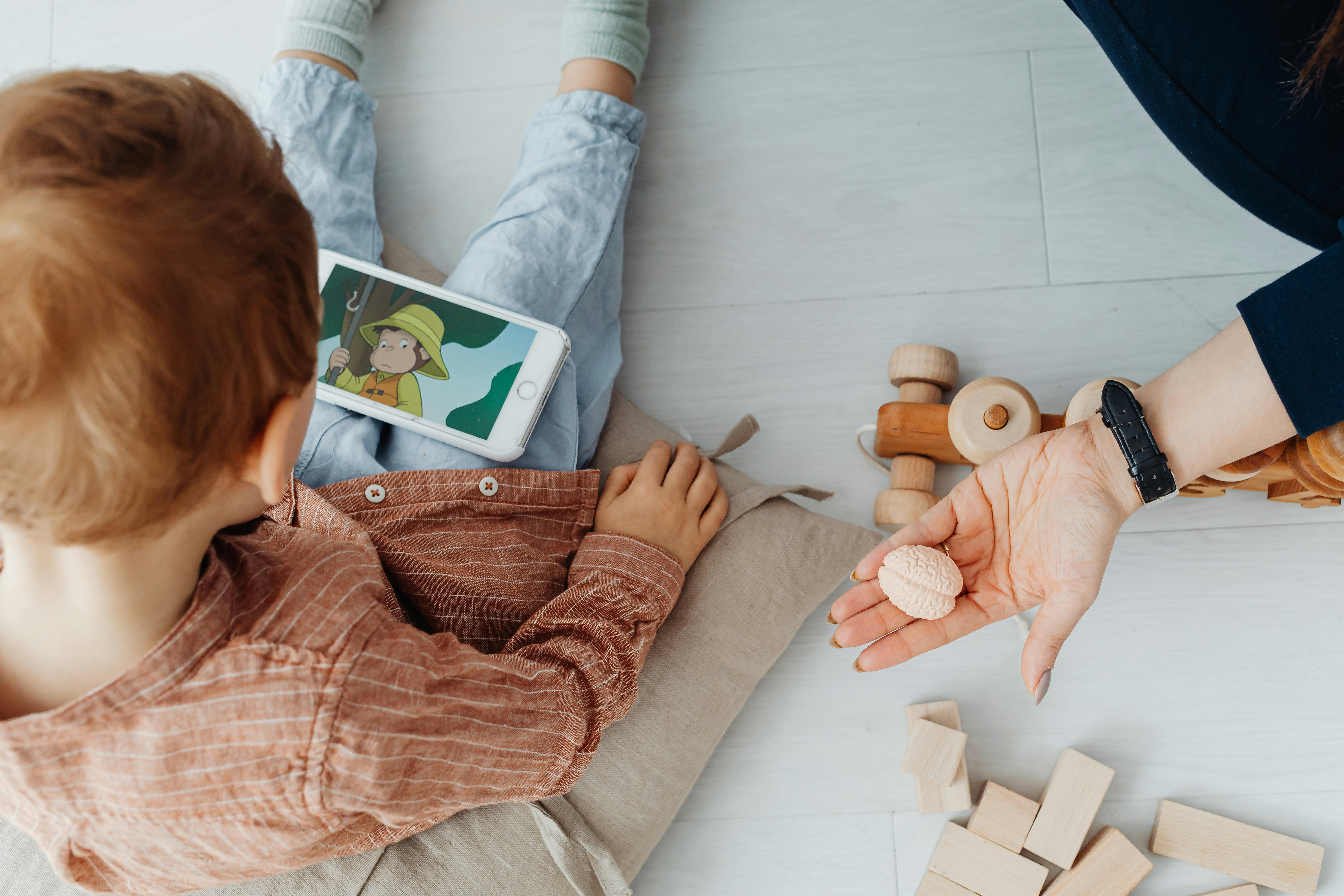 little boy watching a kids show on a smartphone and mother holding a toy in a shape of a little brain