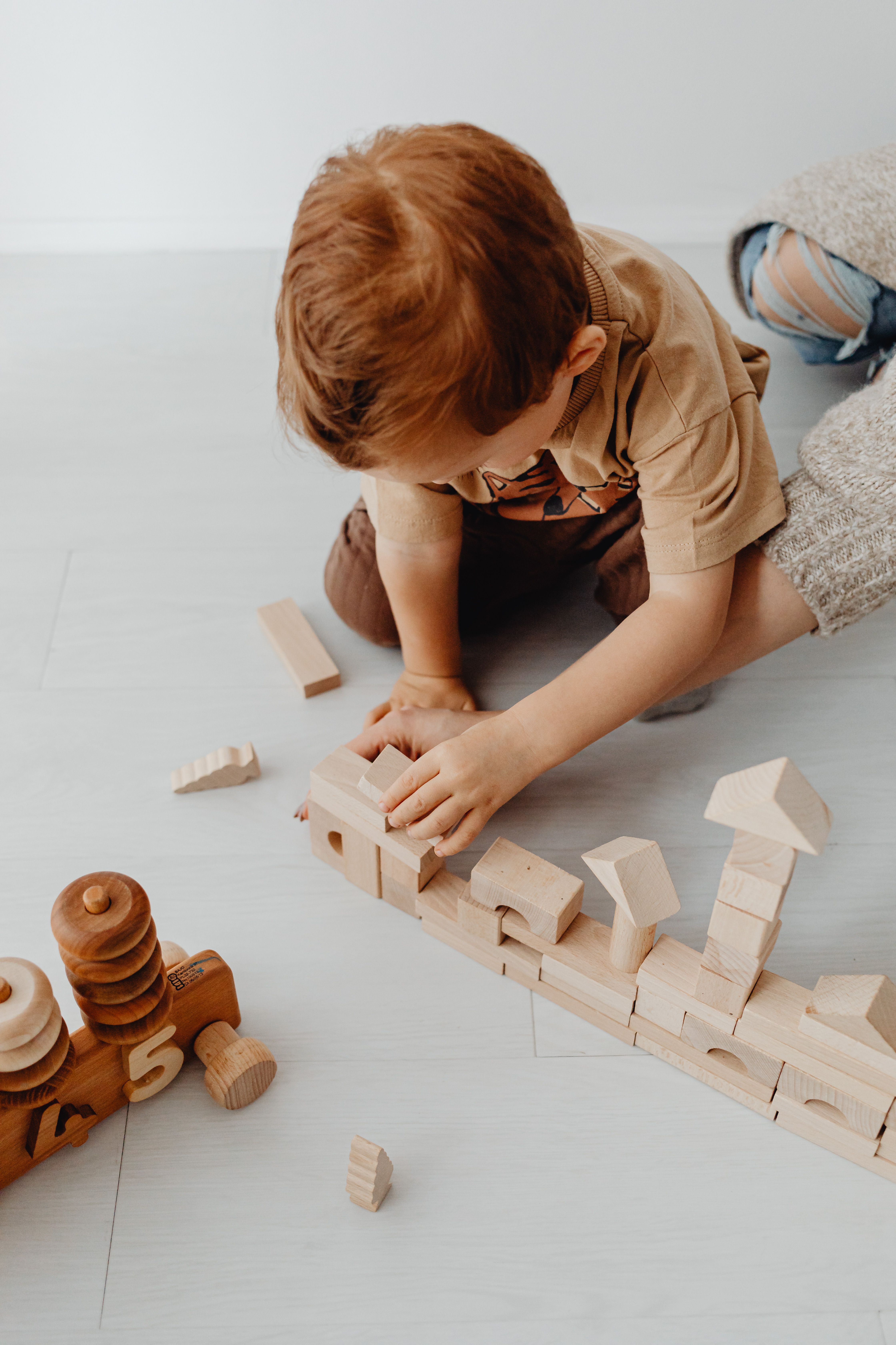 A young child kneels on the floor, absorbed in stacking wooden building blocks into a long row, learning through play.