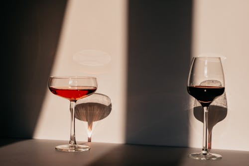 Red wine and vermouth in fragile glasses of various shapes arranged on table near white wall in sunlight