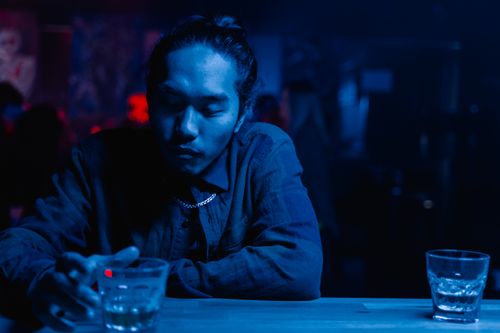 Free Adult man sitting alone in a dimly lit bar, under blue neon lights, holding a cocktail glass. Stock Photo