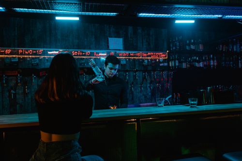 Free A bartender pours a drink under neon lights in a moody bar setting, with a patron seated at the counter. Stock Photo