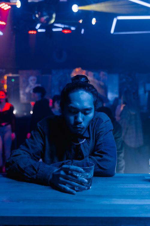 Free An adult man sits at a bar holding a drink, surrounded by a dimly lit, vibrant nightlife scene. Stock Photo
