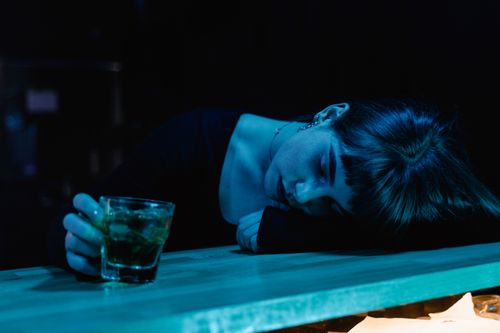 Free A woman resting her head on a bar table while holding a drink, in a moody, dimly-lit nightclub setting. Stock Photo