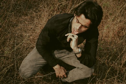 From above of ethnic male in chain and wristwatch pulling back lip while squatting on meadow