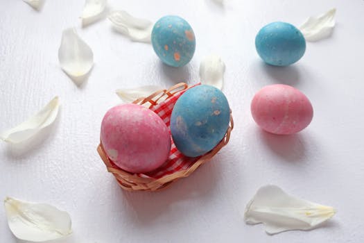 Decorative pink and blue Easter eggs in a small basket surrounded by white petals on a flat surface.