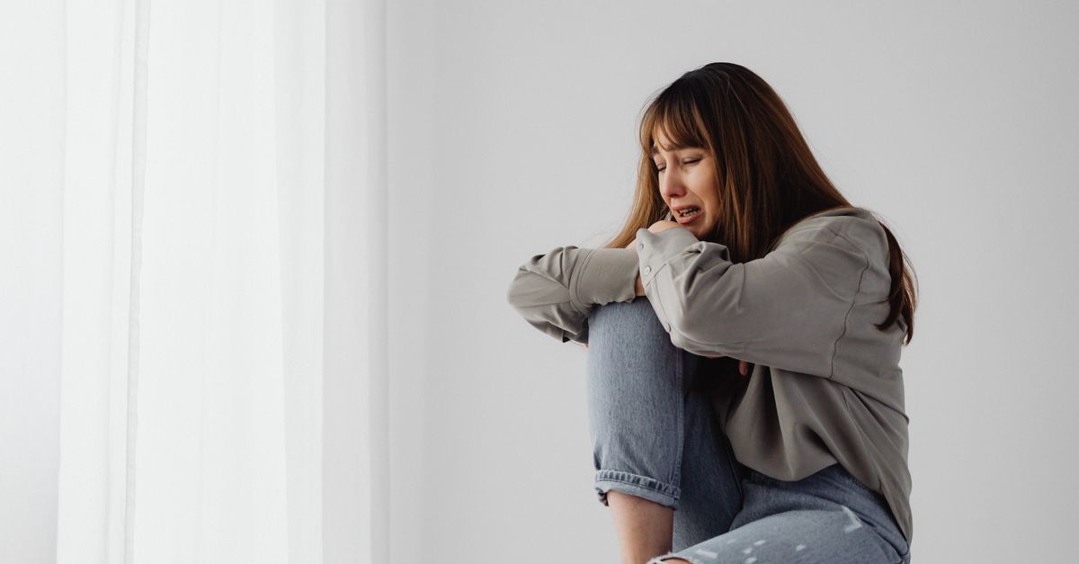 A woman in jeans sits against a white curtain, expressing emotion.