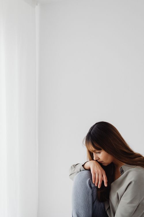 Free A contemplative woman sits near a white wall, looking down with hand on knee. Stock Photo