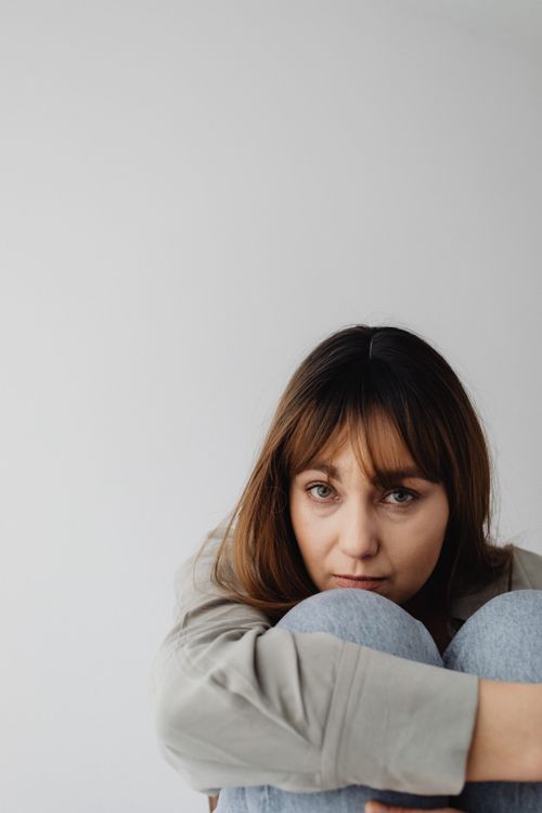 Free Close-up of a woman seated with arms around knees, thoughtful against a gray background. Stock Photo