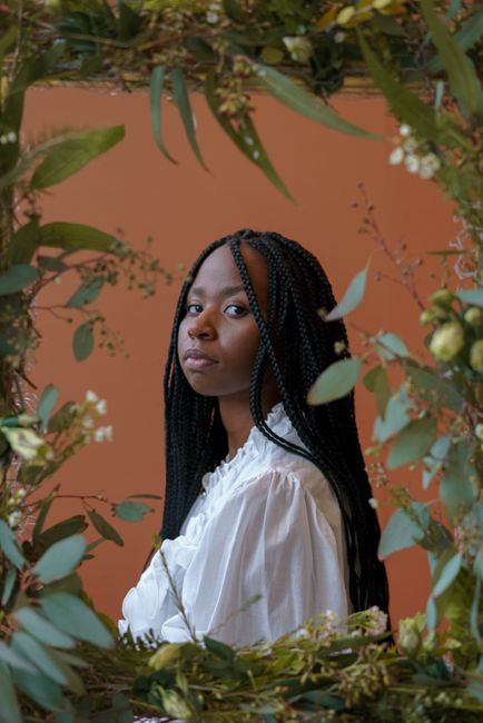 Portrait of young African American woman with braided hair wearing white shirt and looking at camera in frame of green plants