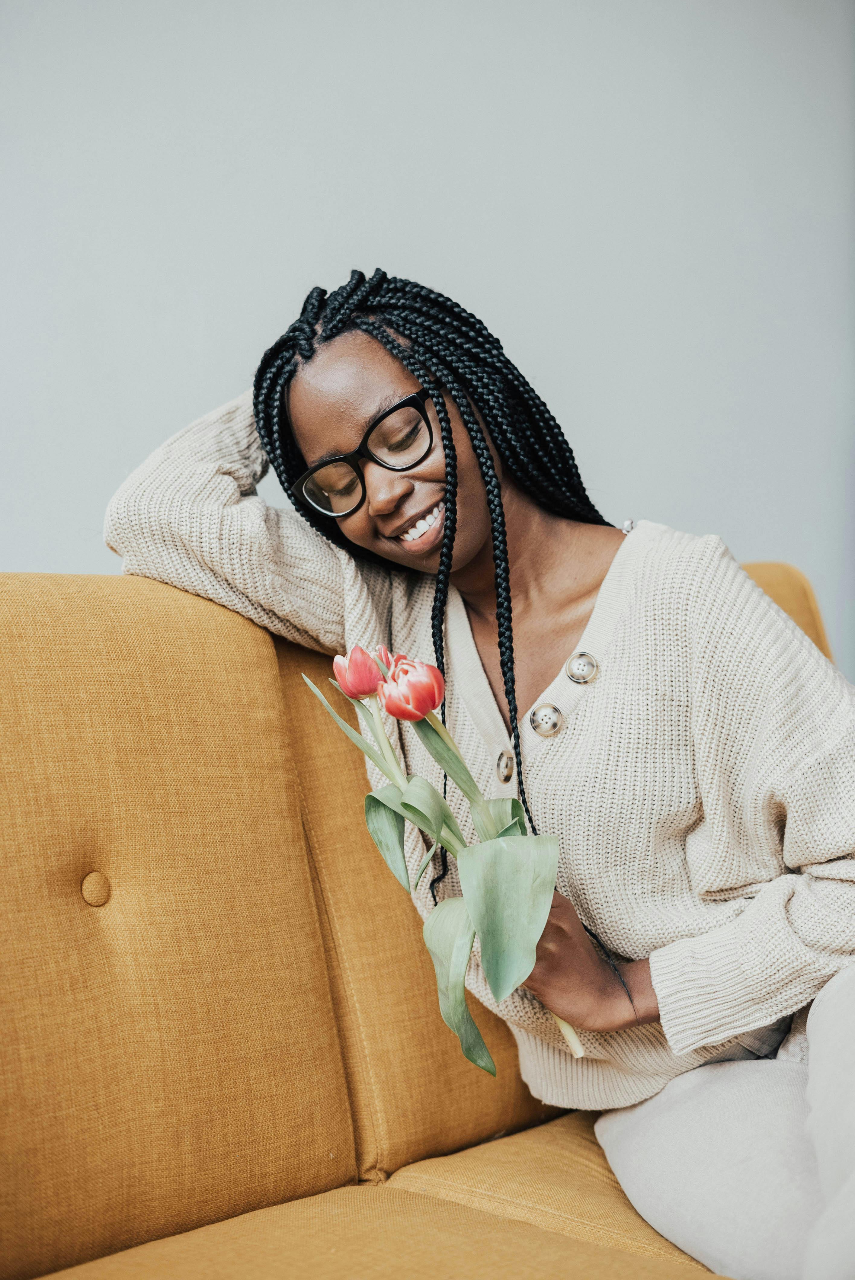 Cheerful African American female in glasses sitting on sofa and looking at tulips dreamily