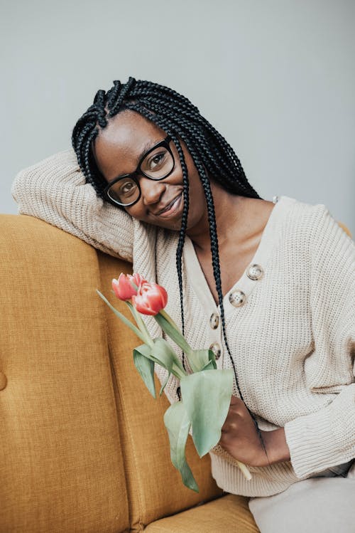 Positive young black female in casual clothes with eyeglasses with tulips with leaves in hand sitting on sofa in light living room while looking away