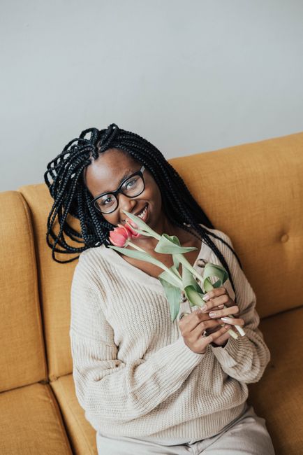 Smiling young African American female in casual outfit with eyeglasses sitting on couch with red tulips with green leaves in hands while looking at camera in bright house