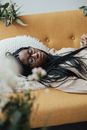 Serious young black female in casual clothes with eyeglasses sleeping on pillow on sofa while lying with closed eyes near green plants and flowers in light living room