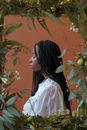 Side view of young serious African American female in white shirt with braids near frame with flowers and plants with green leaves with closed eyes on brown background in studio