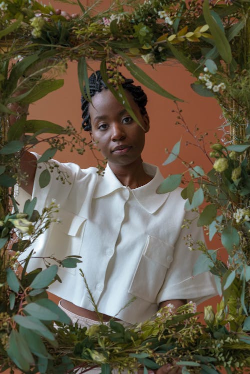 Free Confident young African American lady in white shirt near frame with plants and flowers with green leaves looking at camera in light studio on brown background Stock Photo