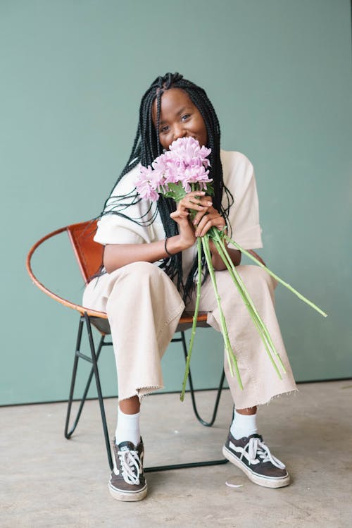 Smiling young African American lady in stylish outfit with cornrows sitting on chair while smelling bouquet of purple flowers while looking at camera in bright room near green wall