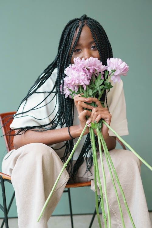 Charming African American female in white clothes sitting on chair with bouquet of flowers in hands and looking at camera