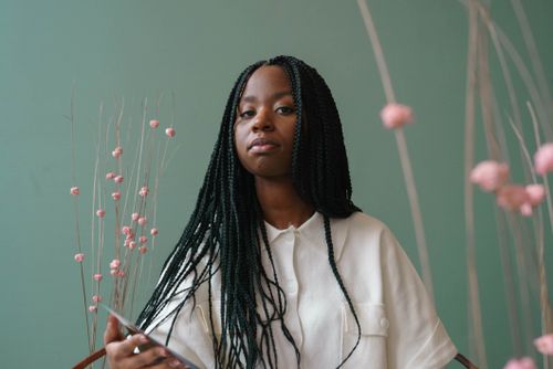 Free Low angle of focused young black female model with long Afro braids in white shirt sitting on chair near decorative plants and looking at camera against green background Stock Photo
