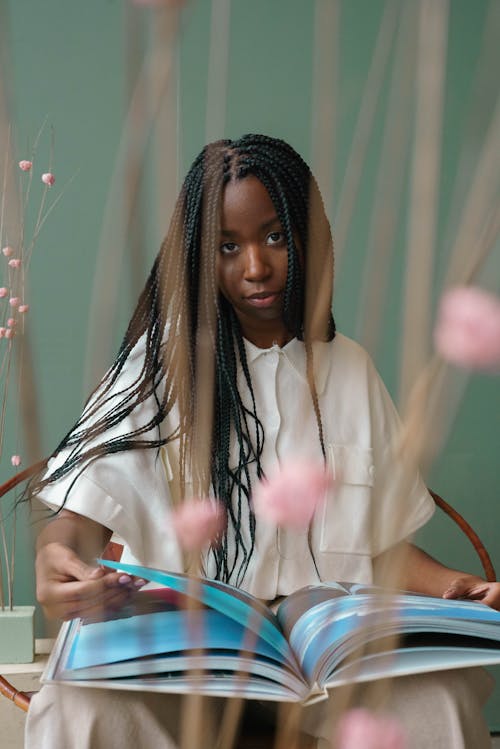 Self assured of young African American female millennial with long dark braids in stylish outfit sitting on chair and reading interesting illustrated book