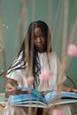 Self assured of young African American female millennial with long dark braids in stylish outfit sitting on chair and reading interesting illustrated book
