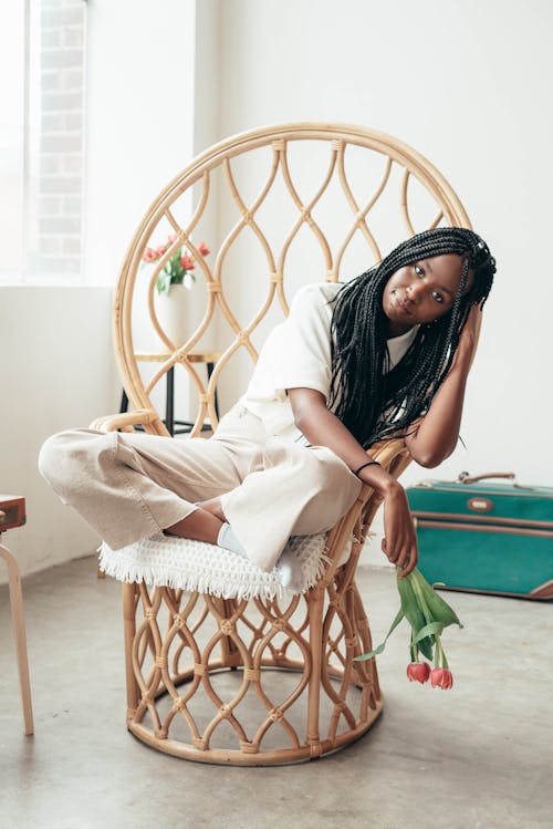 Free Full body of positive young African American lady with long dark hair in casual clothes sitting on stylish bamboo chair with crossed legs and looking at camera Stock Photo