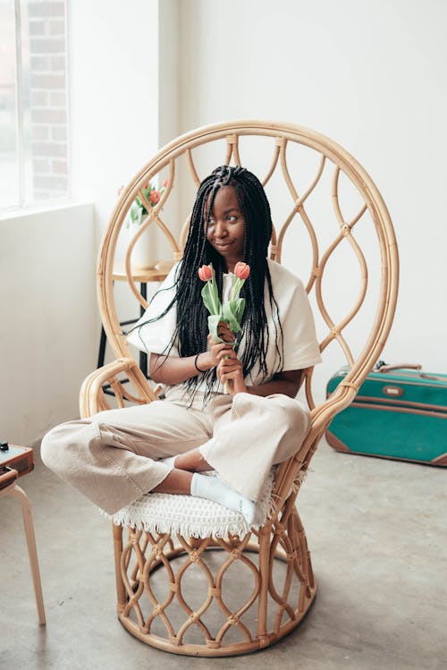 Full body of positive young ethnic lady with long Afro braids holding bunch of fresh tulips and looking away happily while resting in bamboo chair