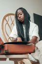 From below of calm young black female meloman with long Afro braids turning on vinyl record player while sitting on chair at home