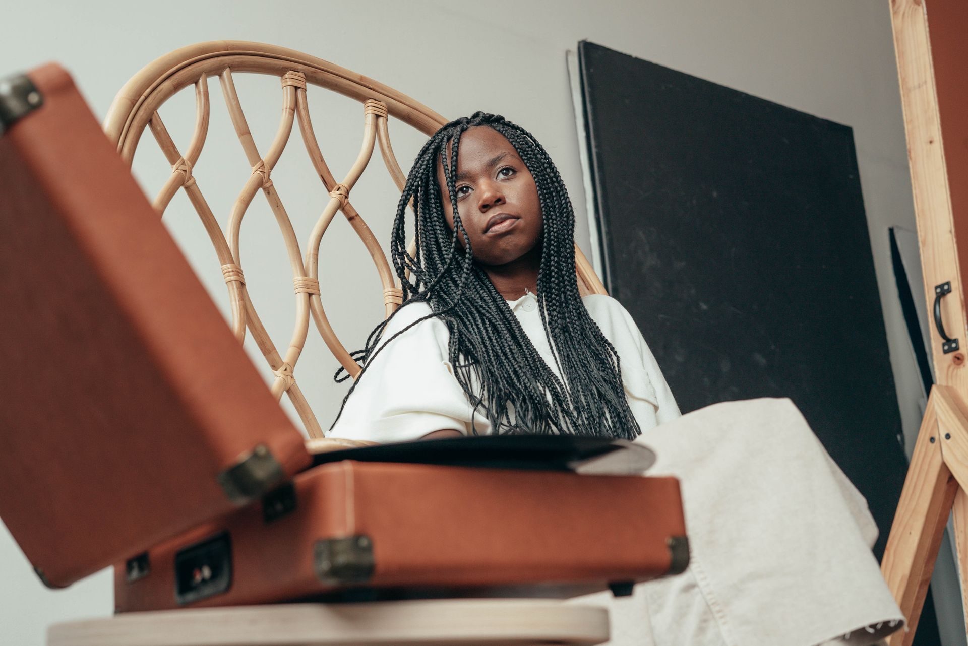 From below of wistful young African American female with long braids listening to music via vinyl record and looking away pensively while resting in bamboo armchair at home