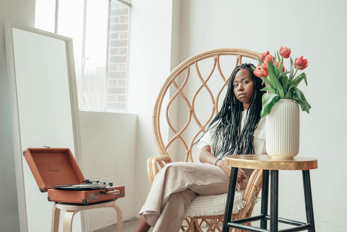 Free Ethnic woman sitting in armchair near vinyl record player and vase of fresh flowers Stock Photo