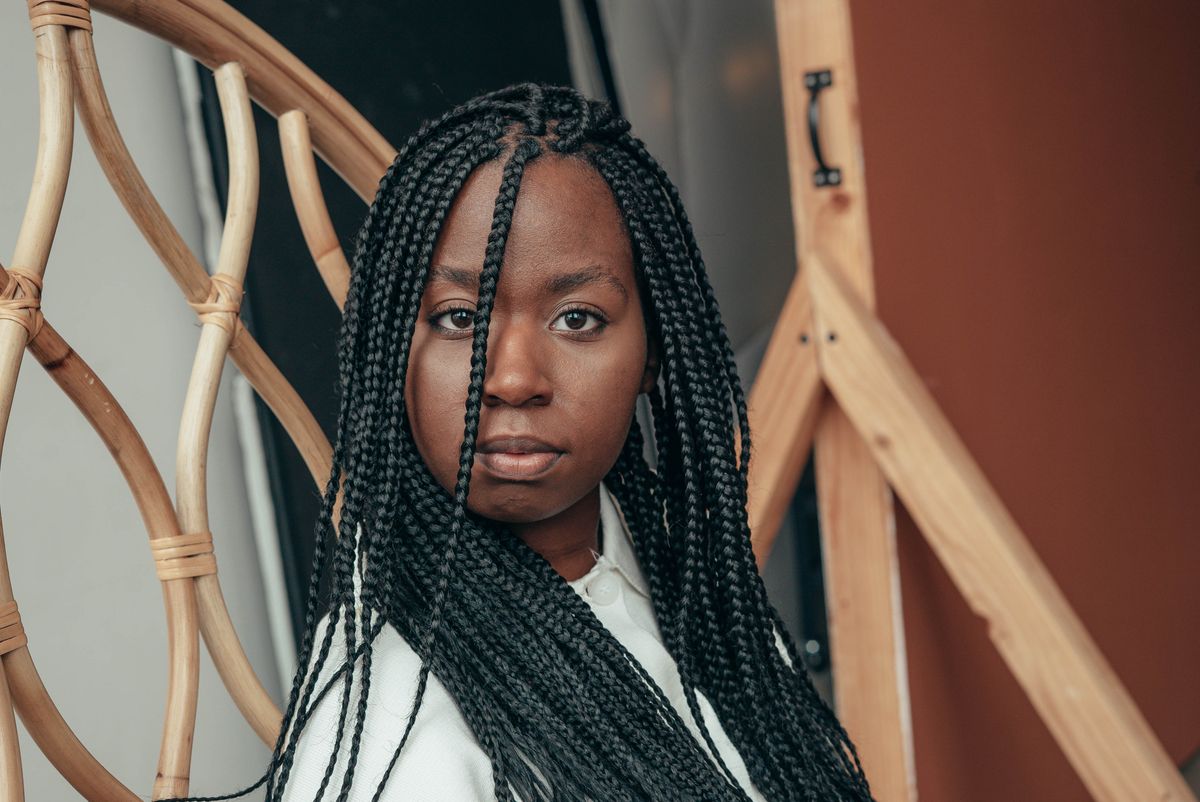 A young Black woman with long box braids and a white collared shirt sits alone in a rattan chair indoors, looking directly at the camera with a calm, level expression.