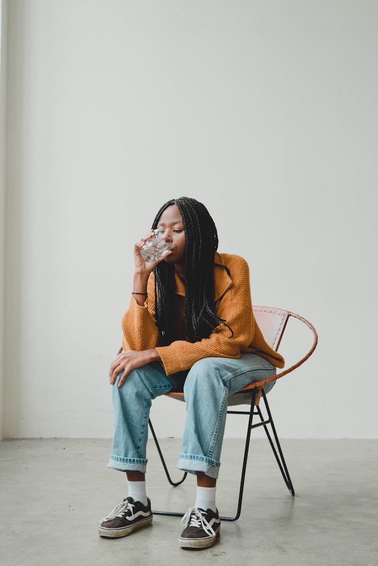 Fashionable Young Black Woman Drinking Water Sitting On Chair