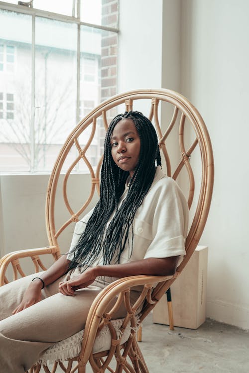 Relaxed young black woman with long Afro braids in casual garment sitting in comfy armchair and looking at camera during weekend at home