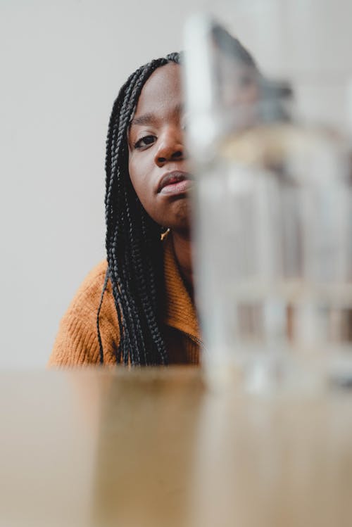 Low angle of serious young African American female millennial with long braids looking at camera through glass of water placed on wooden table