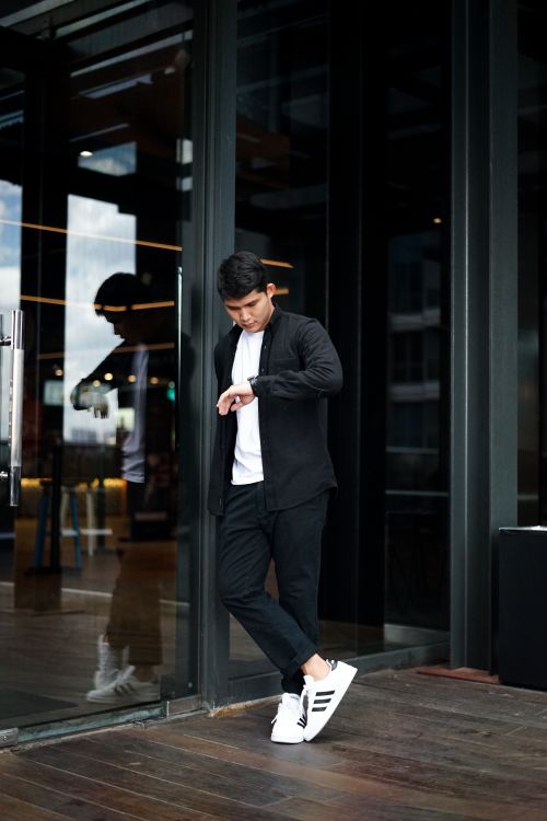 Free Fashionable young man in black outfit checking time on watch outside a modern glass building. Stock Photo