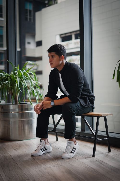 Free A young man in black casual wear sits indoors on a wooden bench with plants nearby. Stock Photo