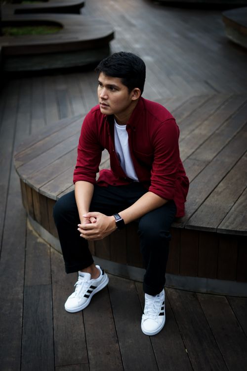 Free A young man in casual attire sits thoughtfully on a wooden platform, showcasing a modern urban style. Stock Photo