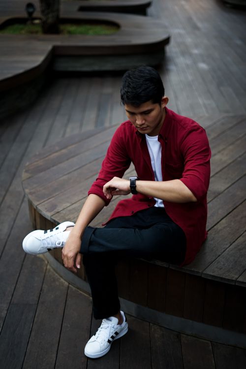 Free Young man in casual attire sitting outdoors, checking his wristwatch on a modern wooden deck. Stock Photo