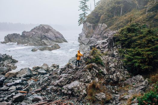A hiker in a yellow jacket explores a rocky coast with dense forest and ocean view.