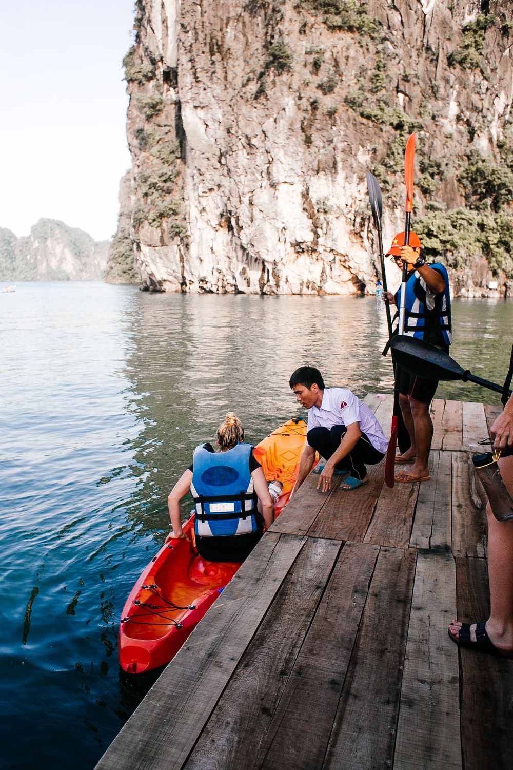 A traveler in a life jacket getting into a kayak with help at a wooden dock