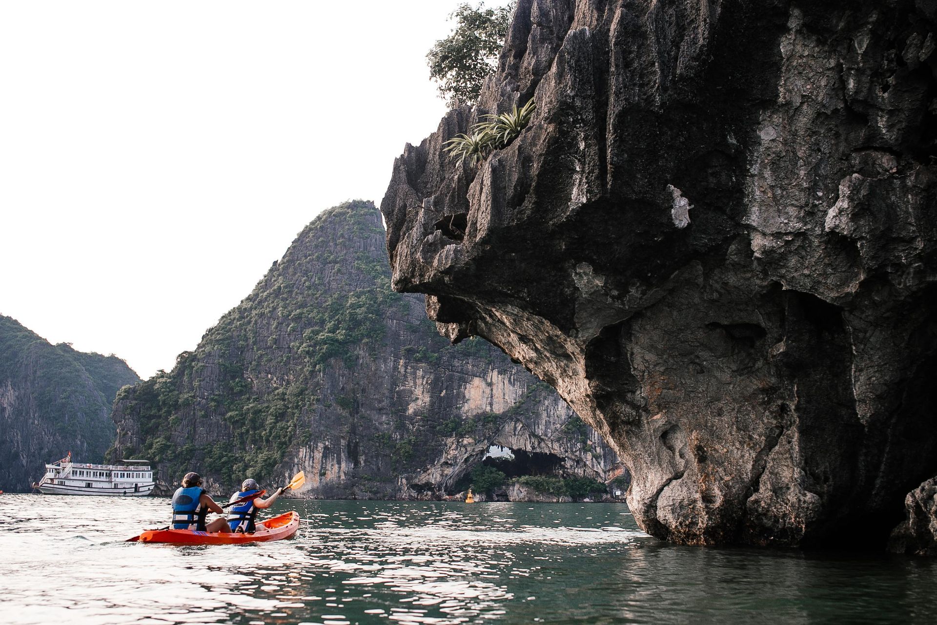 Two kayakers paddle near towering limestone cliffs and a cruise ship.