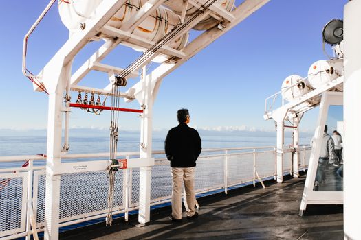 A man stands on a ferry deck admiring the vast sea under a clear blue sky.
