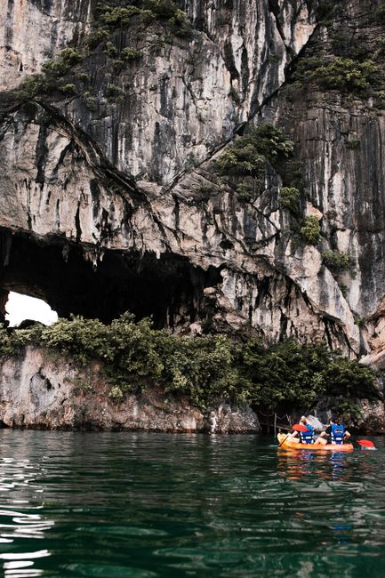 Travelers kayaking in Ha Long Bay near limestone islands and cave entrance, clear green water, bright daylight, relaxed adventure atmosphere, ultra detailed realistic scene