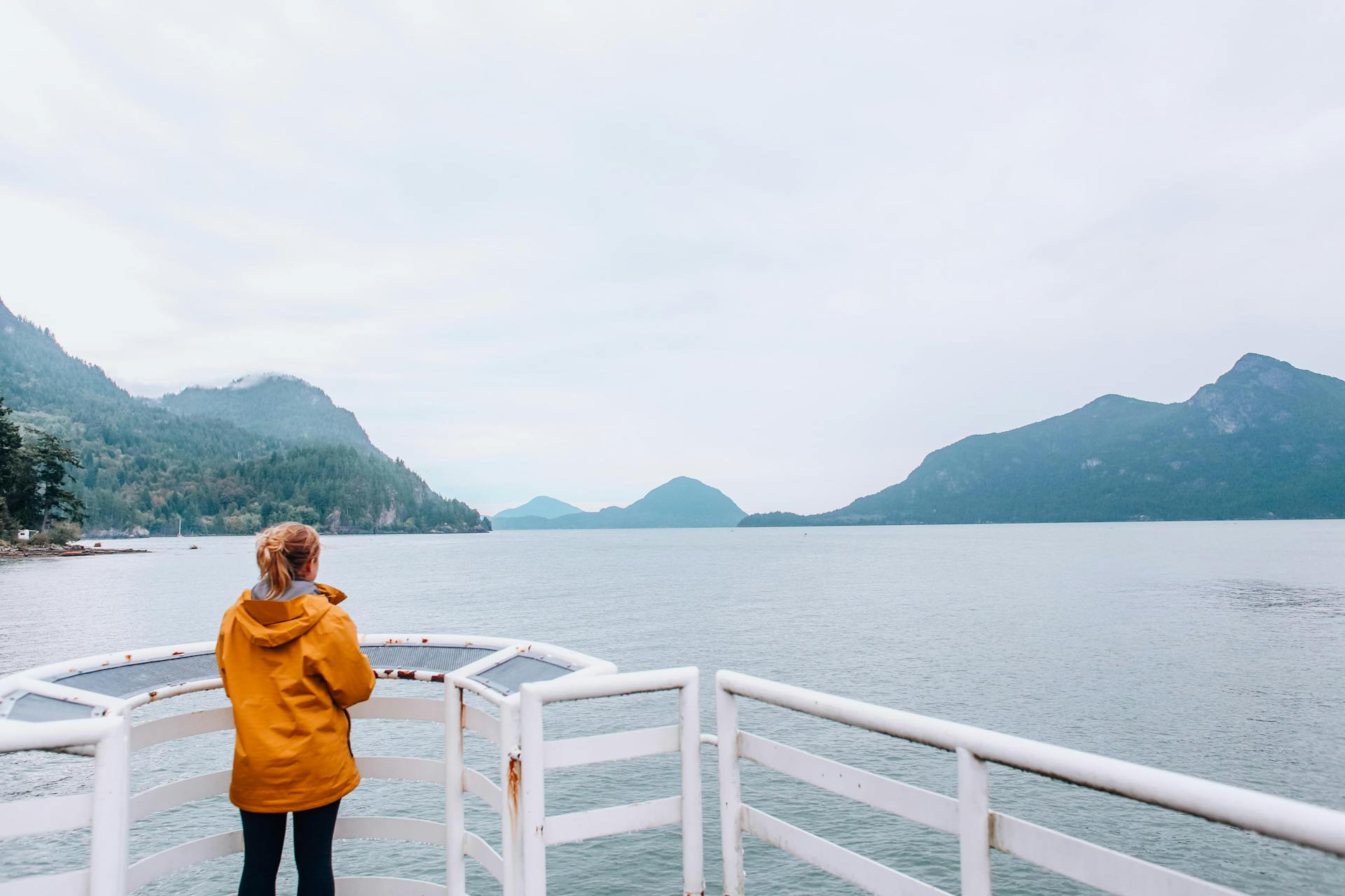Woman stands on a ferry deck, gazing at misty mountains across a serene lake.