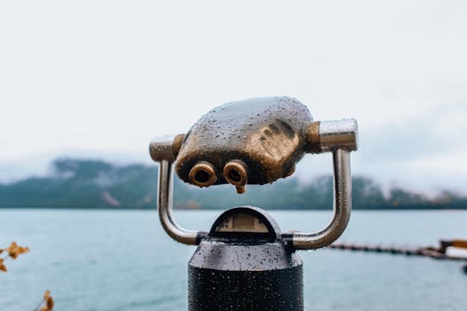 Close-up of a wet coin operated telescope by a lake, capturing a scenic view on a rainy day.